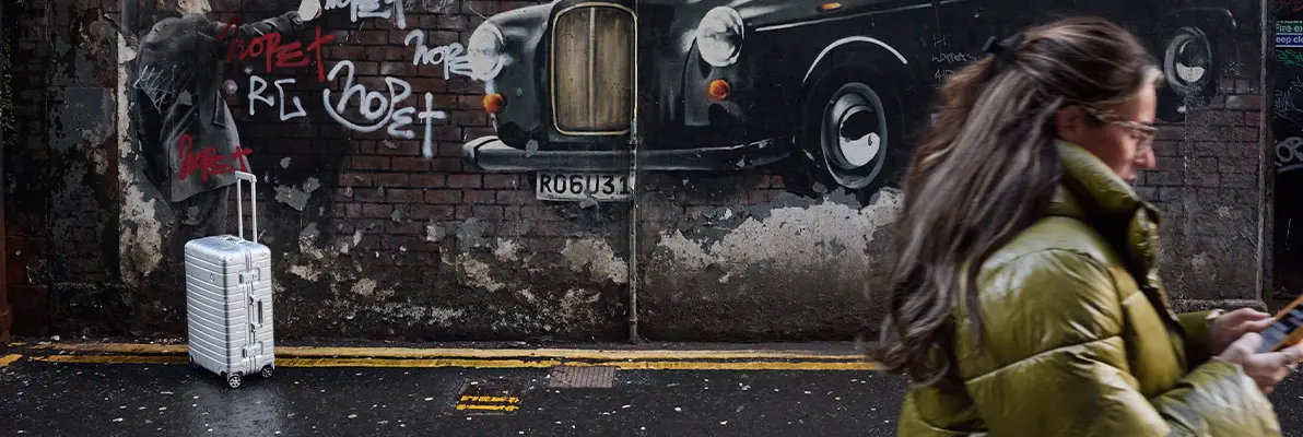 A suitcase from the Lufthansa Aluminium Collection standing against a graffiti-covered wall with a woman passing by in the foreground.