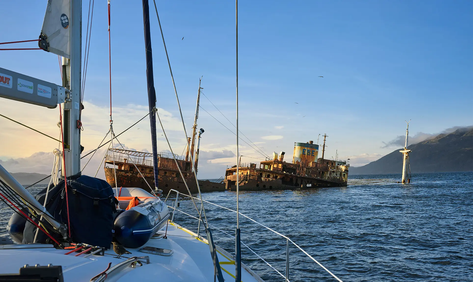 Rusty shipwreck rises from the sea in front of a sailing boat
