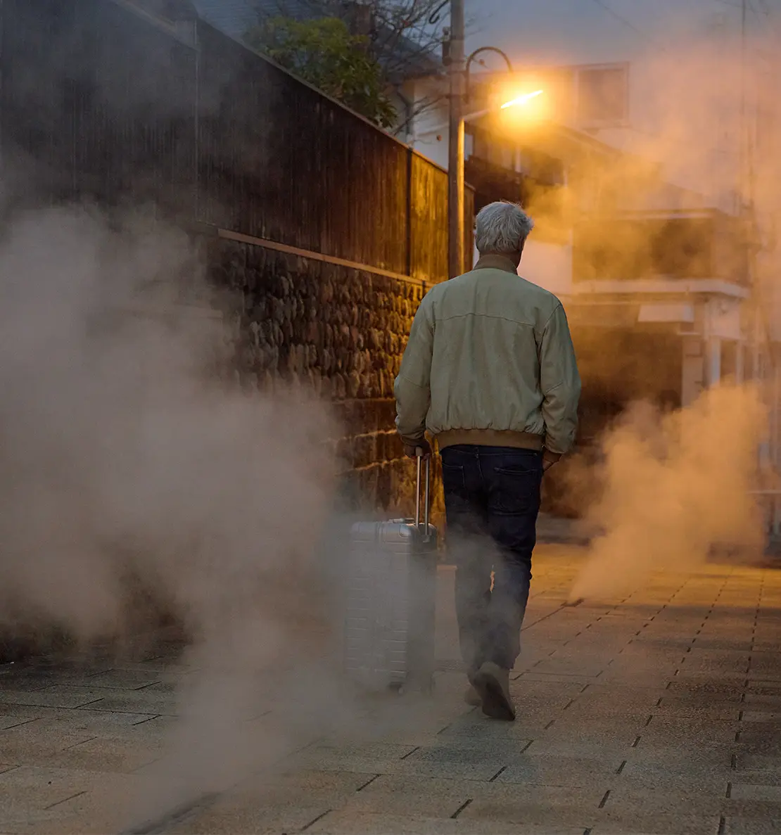 Man with an aluminium suitcase walking through an illuminated alley with volcanic steam rising from the pavement