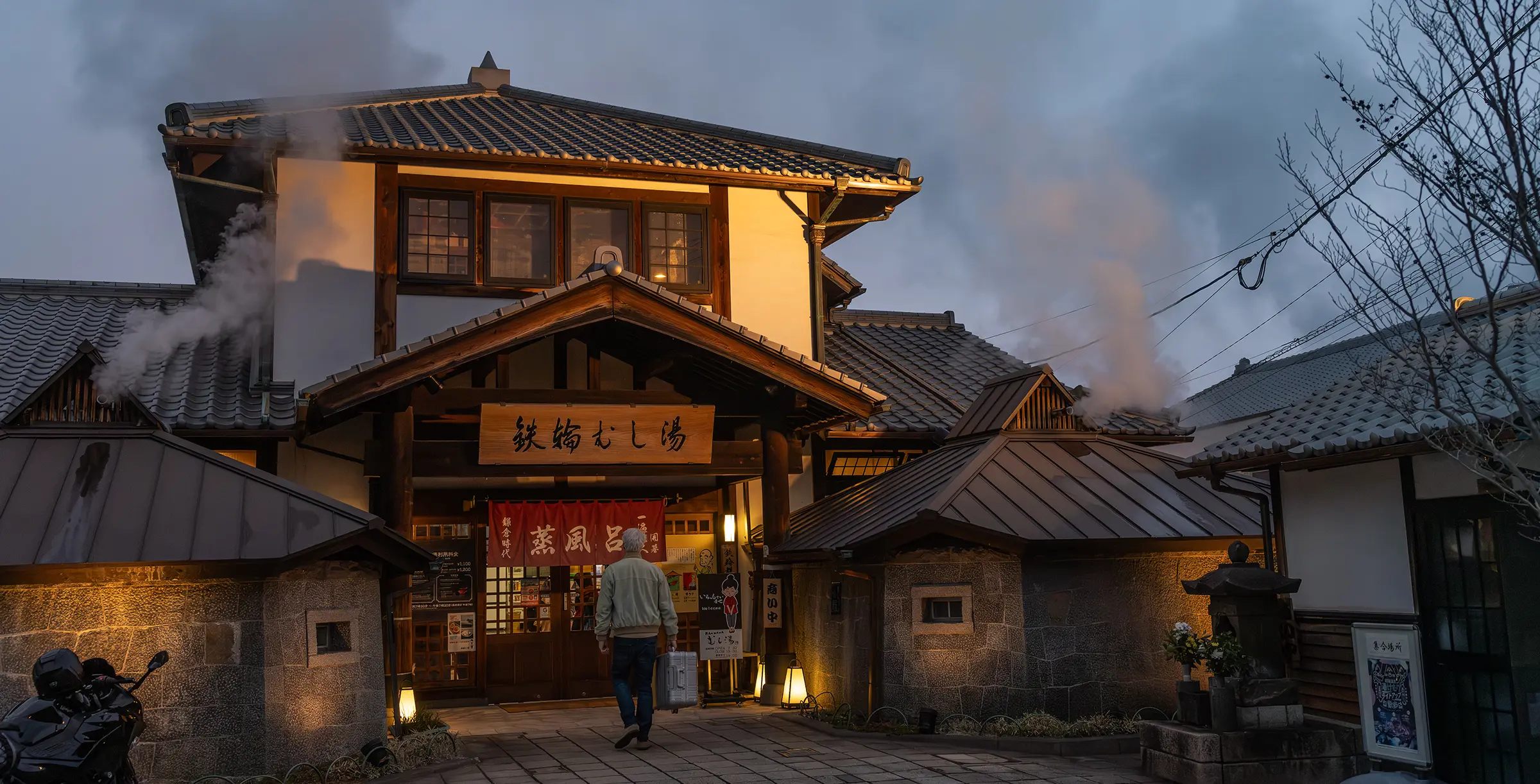 Man with an aluminium suitcase in front of an illuminated onsen bathhouse in the evening – cultural highlight of a trip to Japan