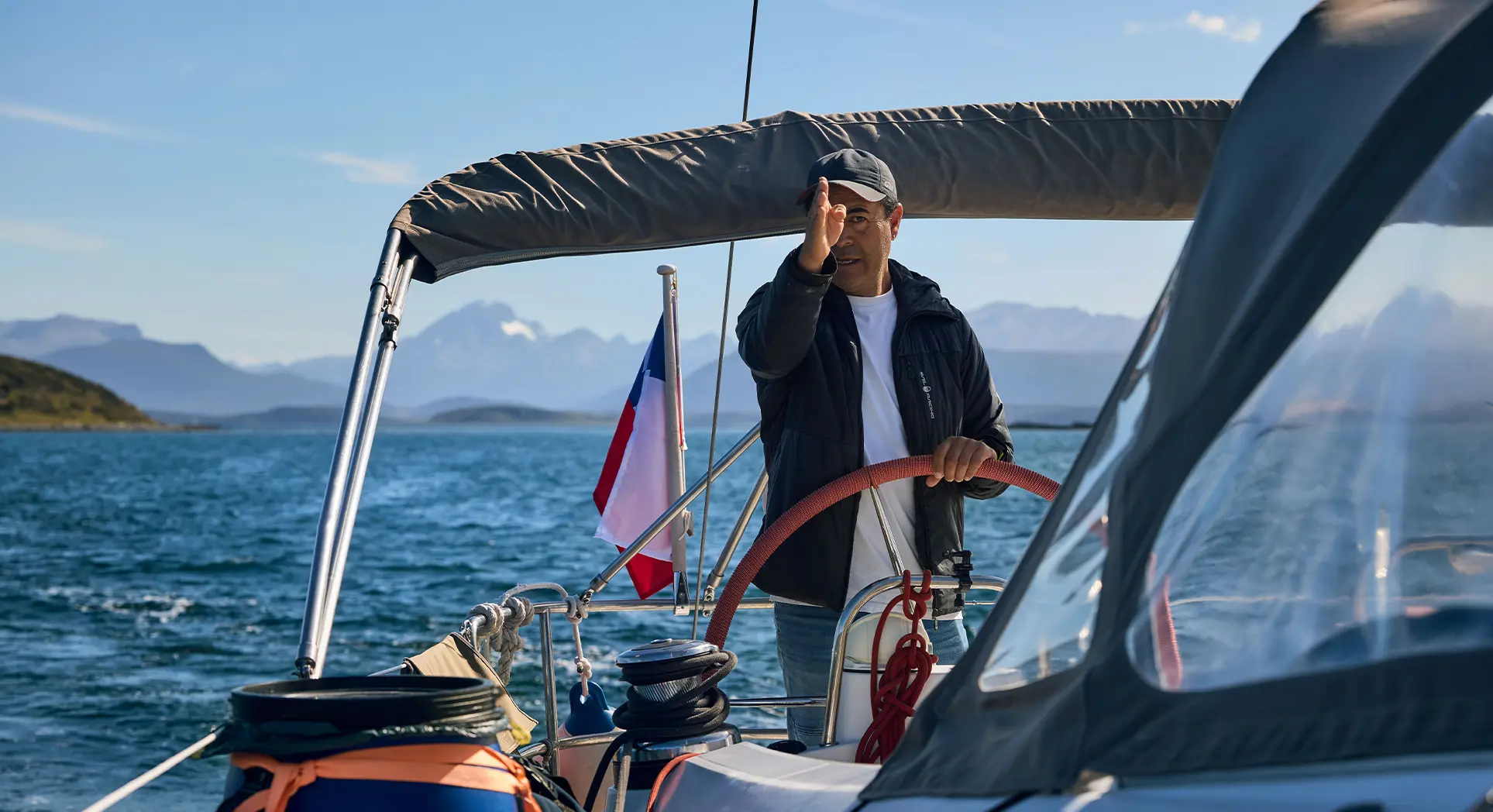 A man stands at the helm of a sailing yacht with a Chilean flag