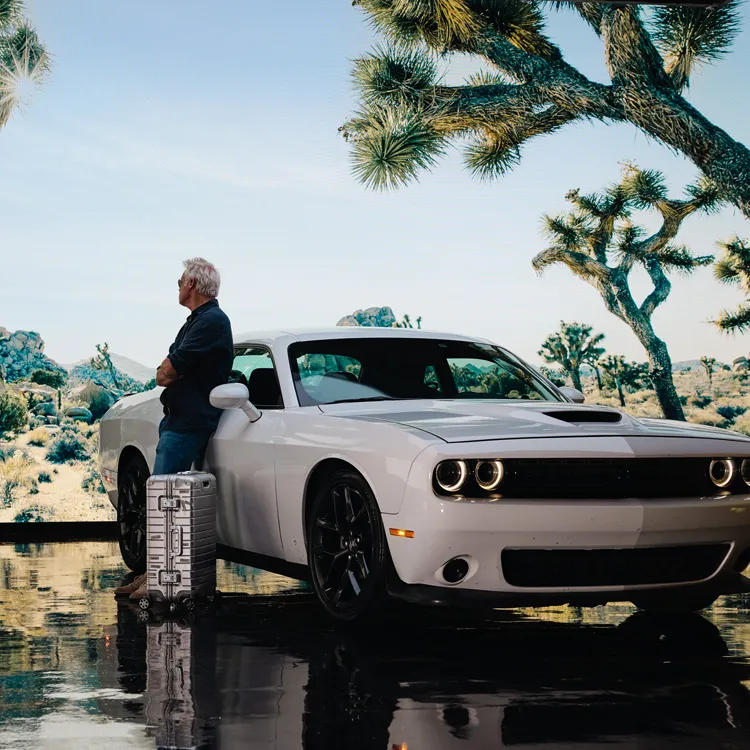 Man with suitcase leaning against Dodge Challenger, desert landscape in background